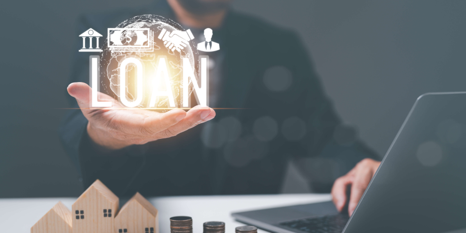 Image of a professional in business attire presenting a holographic projection of the word "LOAN" surrounded by icons of a house, currency notes, handshake, and a person's silhouette. In the foreground, wooden house models and stacked coins on a table suggest real estate financing or mortgage concepts, with a laptop on the side indicating technology integration in banking services.