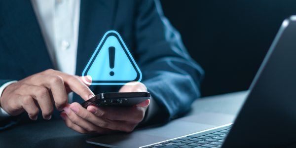 A professional in a suit is using a smartphone in front of a laptop, with a digital exclamation point warning sign appearing above the phone screen, symbolizing a security alert or urgent notification.