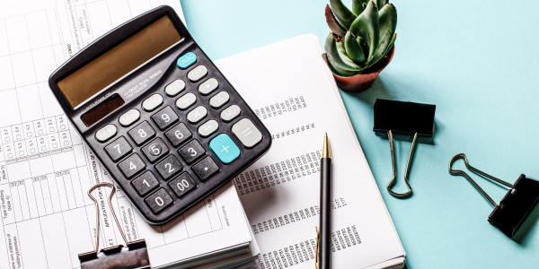 An organized desk setup with a calculator, pencil, binder clips, and documents with tables and text, accompanied by a small green succulent plant, suggesting a professional or academic work environment focused on finance or study.